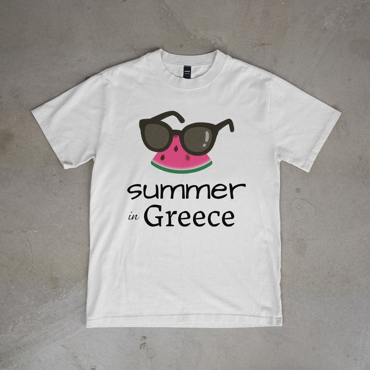 White t-shirt with watermelon and sunglasses graphic and 'summer in Greece' text on a concrete surface.