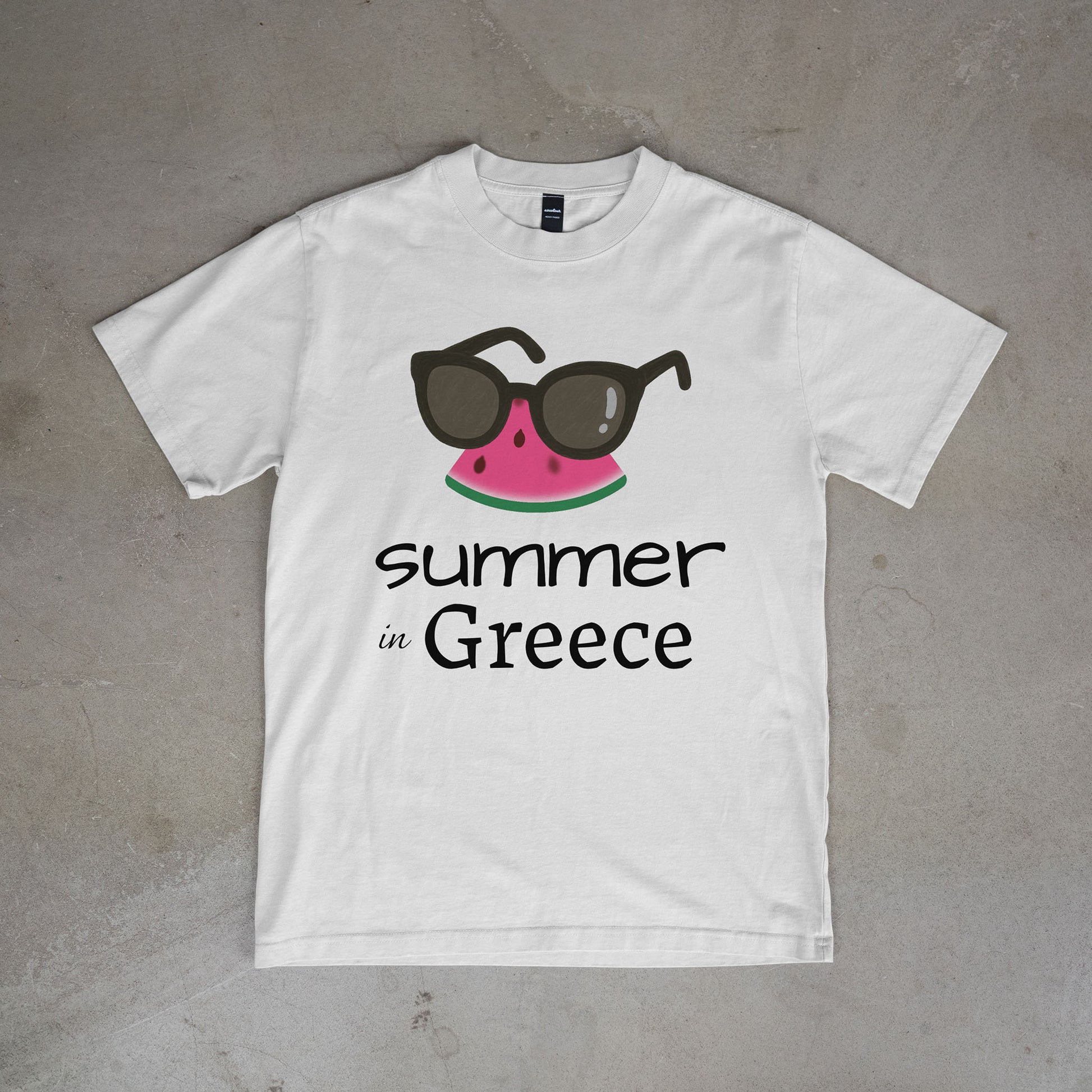 White t-shirt with watermelon and sunglasses graphic and 'summer in Greece' text on a concrete surface.