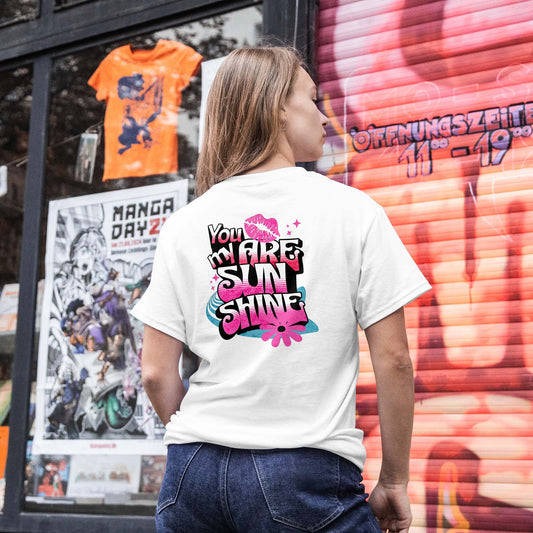 Woman wearing a white t-shirt with colorful text design, standing in front of a store window with posters and a colorful wall.